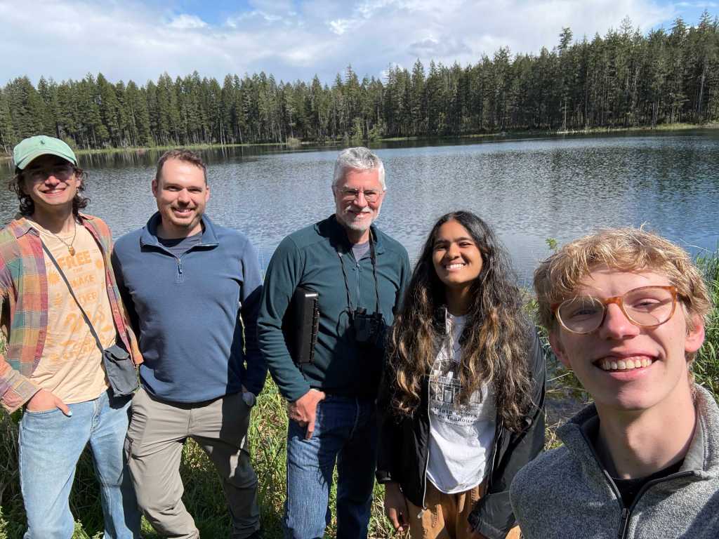 A group of smiling young students surround a bearded middle-aged man in front of a clear lake with an evergreen forest in the background on a mostly sunny day.